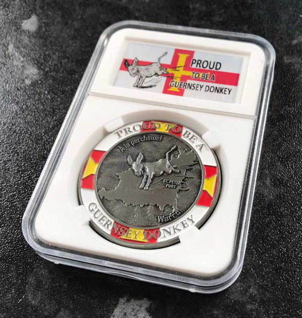 Silver coin with colorful design and 'Proud to be a Guernsey Donkey' text, displayed in a protective case.