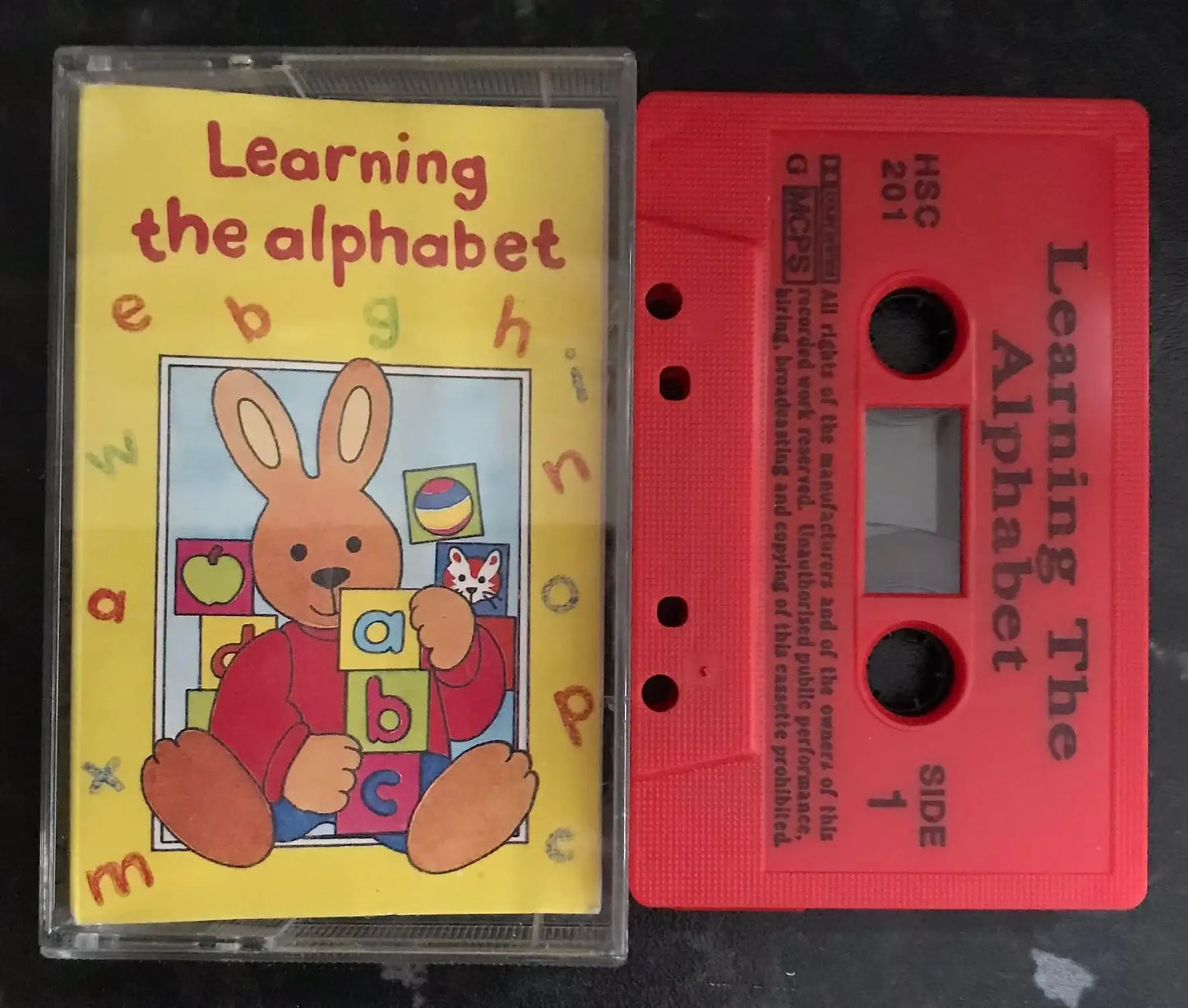 Vintage red cassette tape and yellow album cover for "Learning The Alphabet" with a bunny character.